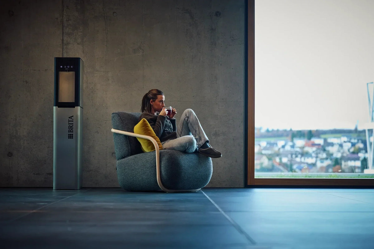 Un distributeur d’eau BRITA dans un environnement de bureau moderne, à côté d’une personne travaillant sur un ordinateur portable assise dans un fauteuil.