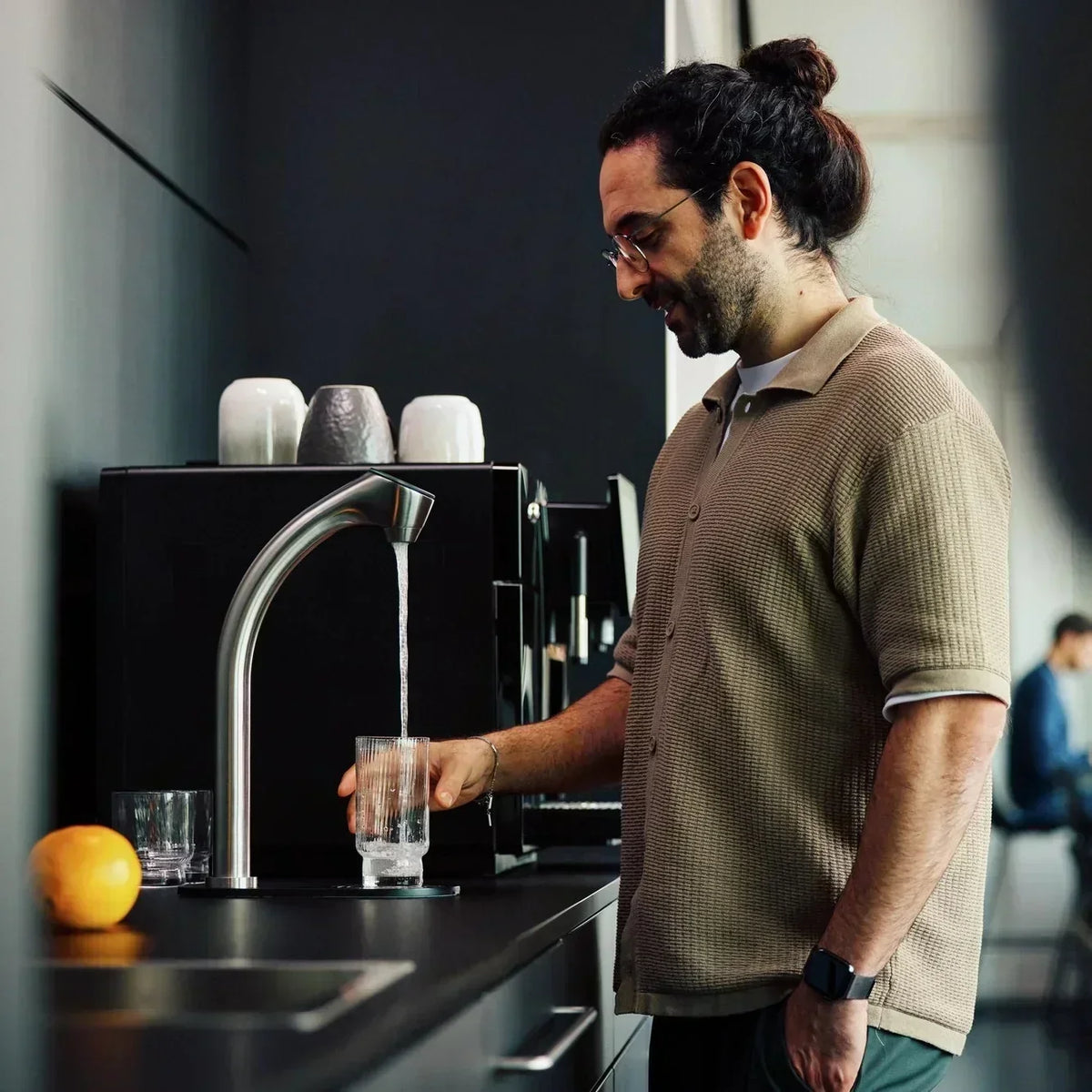 Un homme se tient dans une cuisine et remplit un verre d’eau au robinet.