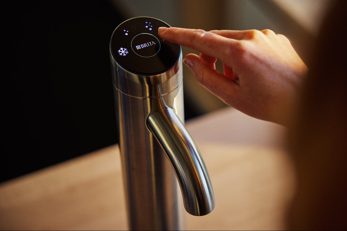 A close-up of a stainless steel water dispenser with a black touch control panel, being operated by a person's finger in a warm indoor setting.
