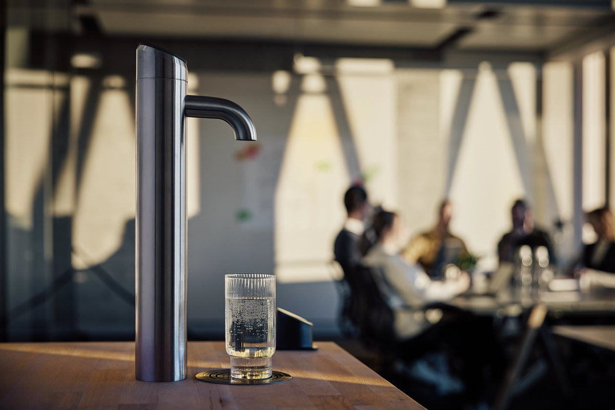 Extra I-Tap dispenser on a wooden table with a glass of water in a blurred office setting.