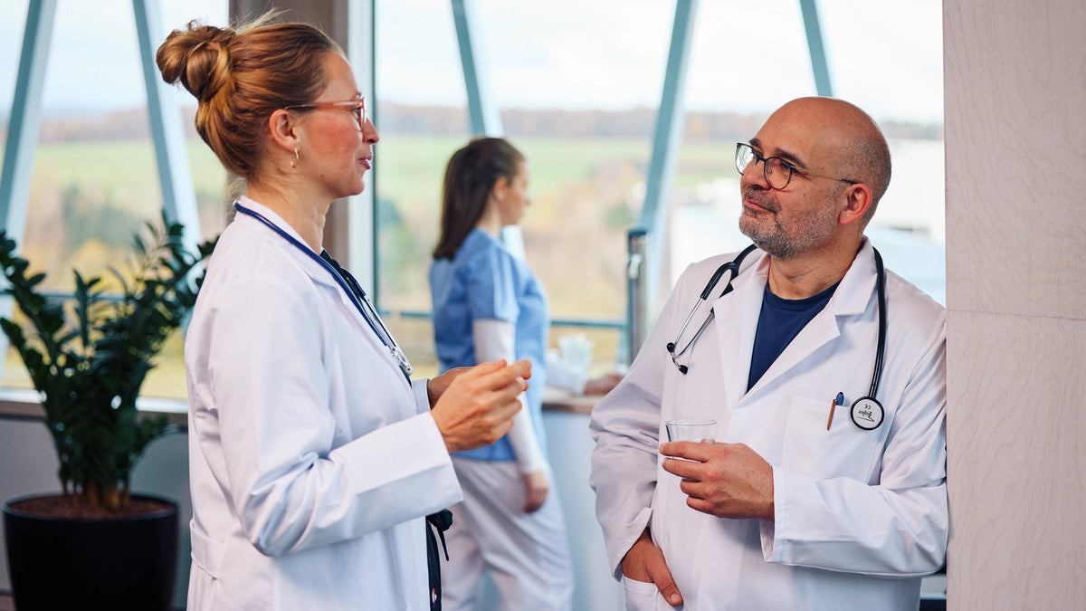 Two doctors in a conversation in a hospital setting, with a person in the background