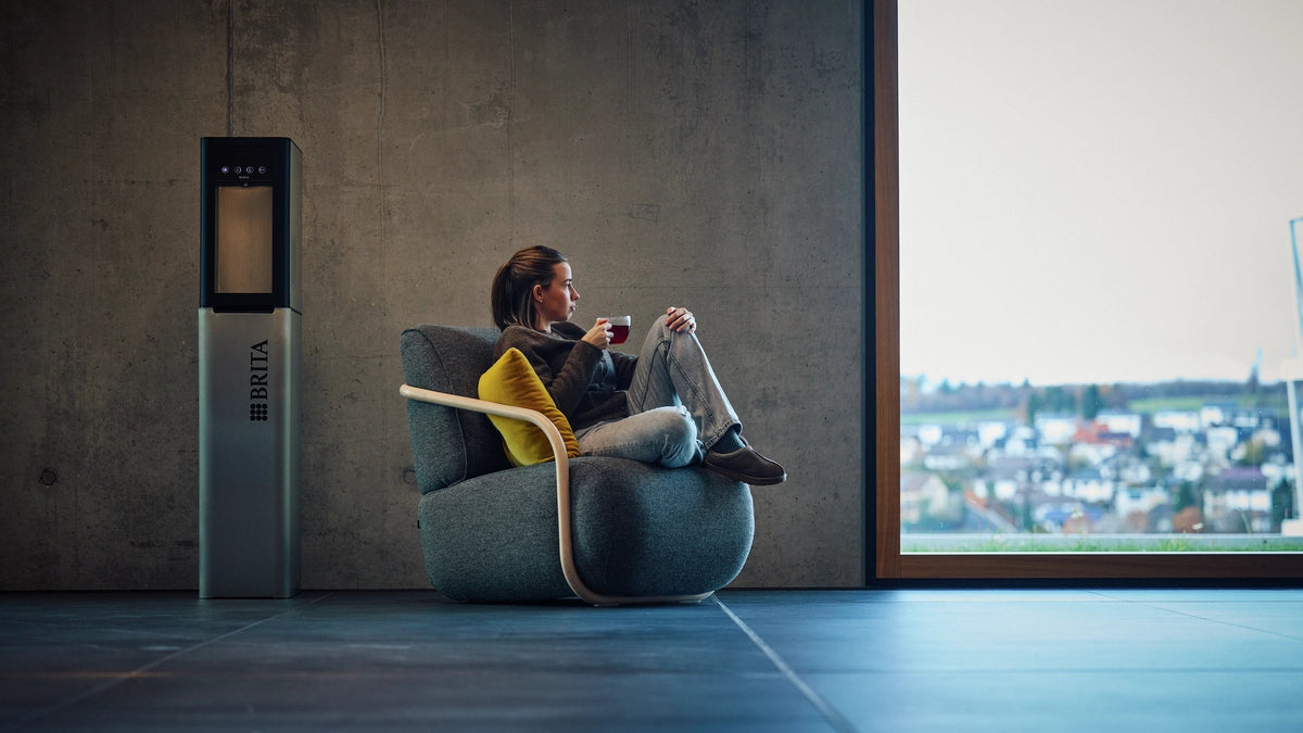 Woman sitting in a modern chair by a window with a view, next to a water dispenser.