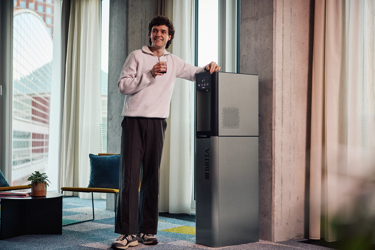 Person standing next to BRITA Top Pro in an indoor setting with large windows, holding a glass of water.