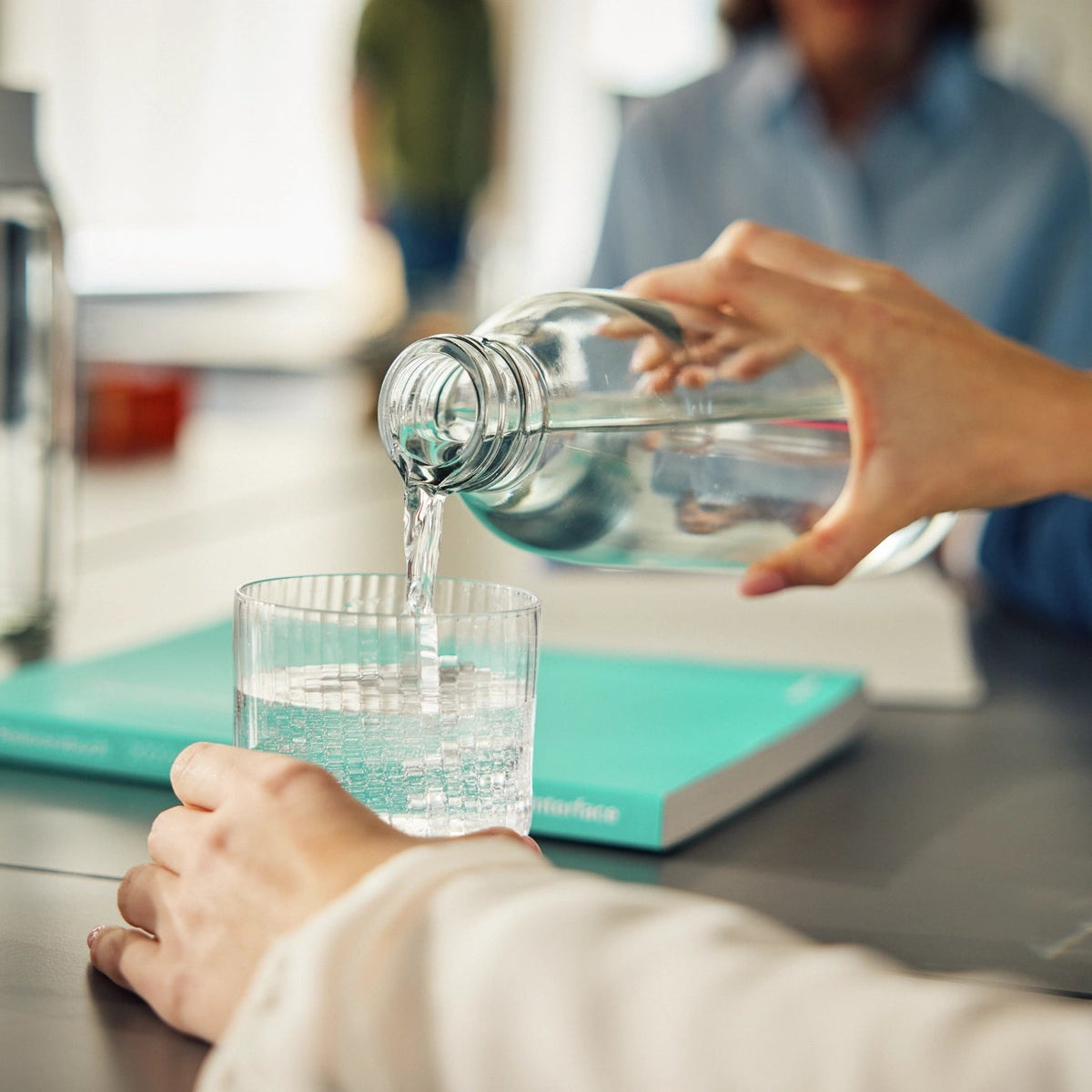 Una persona che versa dell'acqua da una bottiglia in un bicchiere appoggiato su una scrivania.